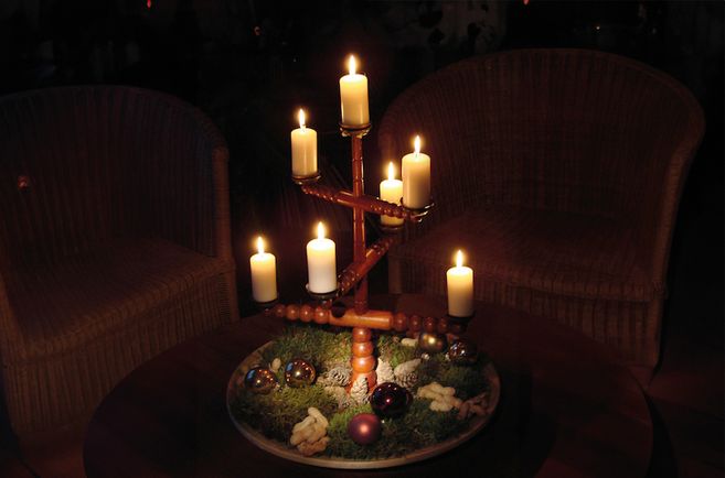 Ein traditioneller Kerzenhalter mit sieben brennenden Kerzen steht auf einem Tisch, umgeben von Moos, Tannenzapfen und Weihnachtskugeln.