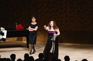 Zwei Frauen auf einer Bühne, eine singt leidenschaftlich, die andere steht mit verschränkten Armen daneben. Ein Flügel im Hintergrund.