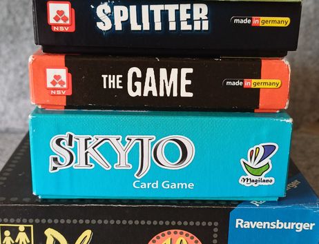 Ein Stapel von Brettspielen, darunter Qwantum, Splitter, The Game, Skyjo und Phase 10, mit mehreren bunten Würfeln davor.