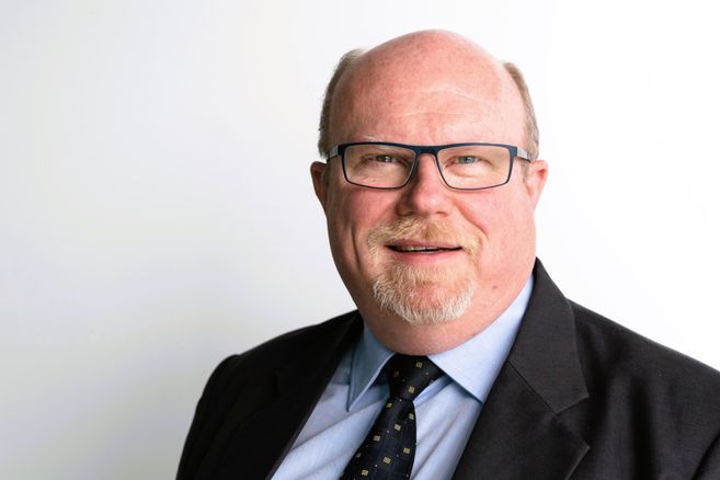 Porträt eines lächelnden Mannes mit Brille und Anzug vor weißem Hintergrund.