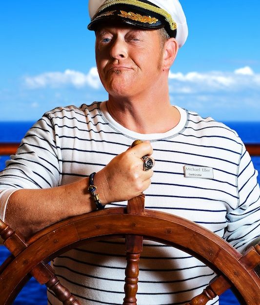 Person in Kapitänsuniform am Steuerrad eines Schiffs.