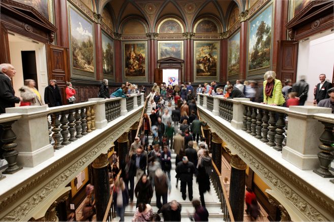 Menschenmenge in einem kunstvoll dekorierten Museum mit Gemälden an den Wänden.