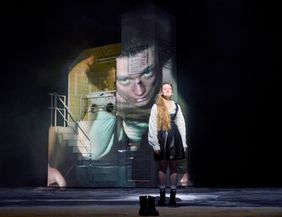 Ein Schauspieler steht auf einer Bühne in historischer Kleidung. Im Hintergrund ist ein großes projiziertes Bild mit einem nachdenklichen Gesicht.