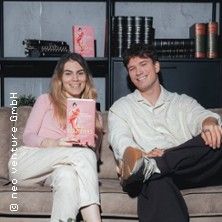 Zwei Personen sitzen auf einem Sofa, eine hält ein Buch mit rosa Cover in der Hand. Im Hintergrund sind Bücherregale zu sehen.