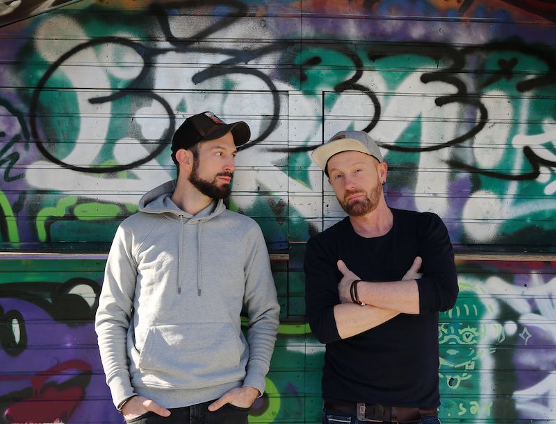 Zwei Männer stehen vor einer mit Graffiti besprühten Wand.