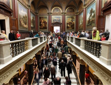 Menschenmenge in einem kunstvoll dekorierten Museum mit Gemälden an den Wänden.
