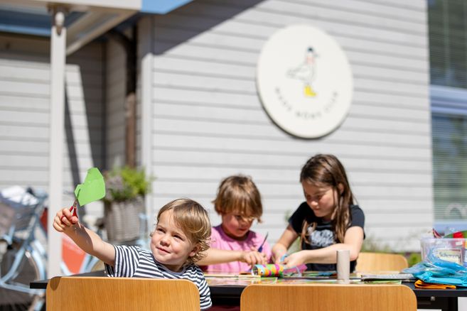 Kinder basteln an einem Tisch im Freien vor einem Gebäude mit einem Schild des Mads Möwe Clubs.