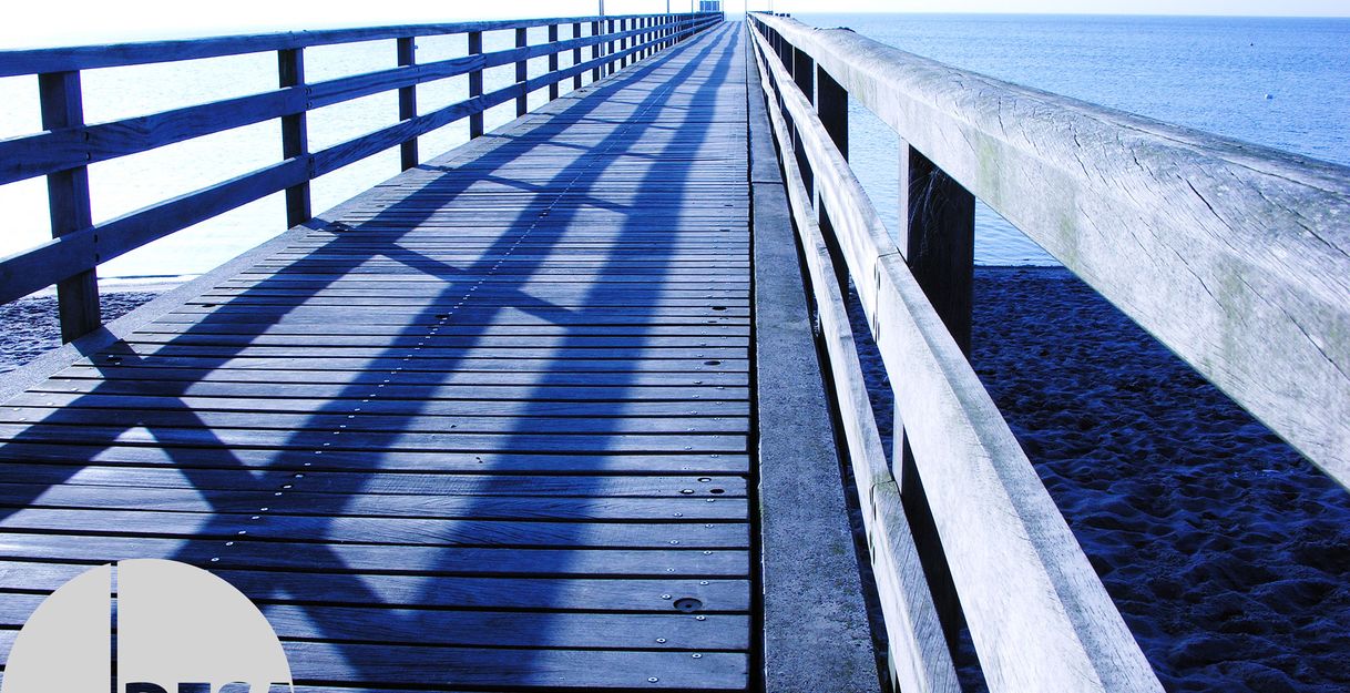 Eine lange Holzseebrücke erstreckt sich ins Meer, beleuchtet von sanftem Morgenlicht. Die Schatten der Geländer fallen diagonal über die Planken.