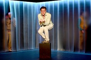 Ein Mann in weißer Kleidung hockt auf einem Podest in einem beleuchteten Raum mit transparenten Vorhängen.