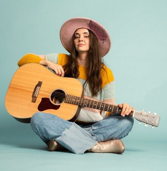 Frau mit Gitarre und Hut sitzt im Schneidersitz vor blauem Hintergrund.