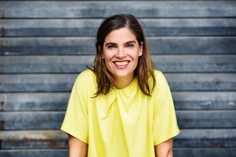 Frau in gelbem T-Shirt lächelt vor einer grauen Holzwand.