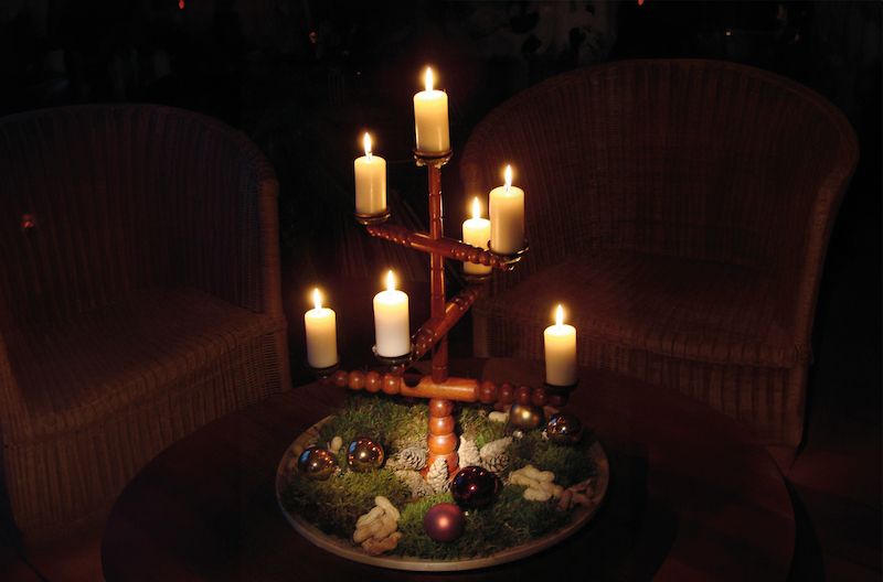 Ein traditioneller Kerzenhalter mit sieben brennenden Kerzen steht auf einem Tisch, umgeben von Moos, Tannenzapfen und Weihnachtskugeln.