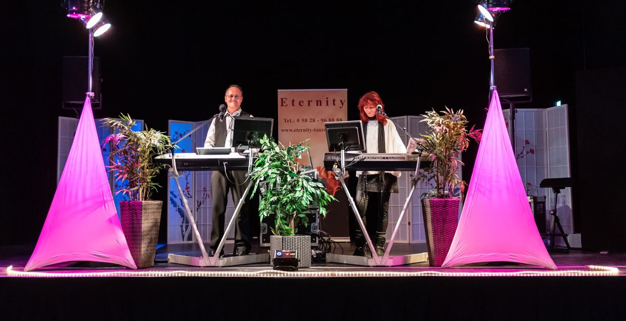 Zwei Musiker auf einer Bühne mit Keyboards und Mikrofonen, umgeben von Pflanzen und pinkem Licht.