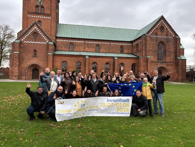 A group of people stands in front of a church holding up a banner. The banner displays information about an event in the city center.