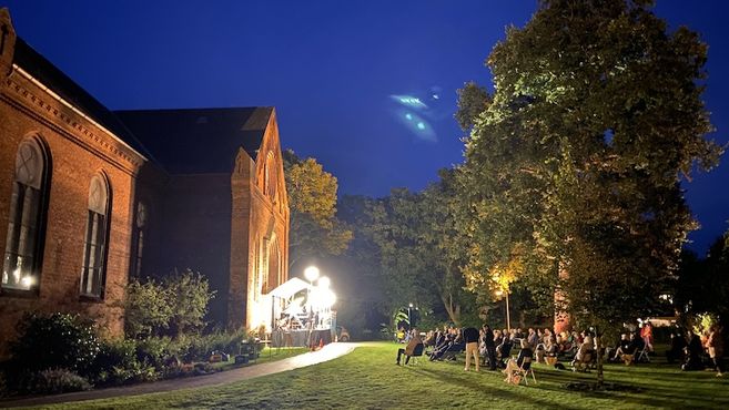 Open-Air-Konzert auf einer Wiese neben einem Backsteingebäude bei Nacht.