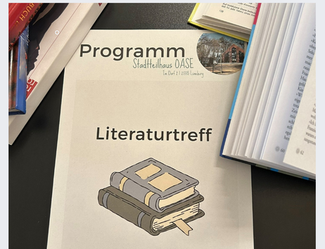Ein Programmheft mit der Aufschrift 'Literaturtreff' liegt auf einem Tisch, umgeben von geöffneten Büchern.