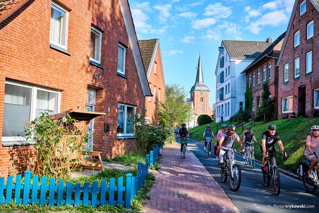 Gruppe von Radfahrern auf einer Straße mit Backsteinhäusern und Kirchturm im Hintergrund.
