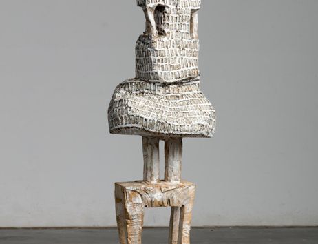 Eine abstrakte Skulptur aus Holz, die eine menschliche Figur darstellt. Sie hat einen runden Kopf und eine strukturierte Oberfläche.