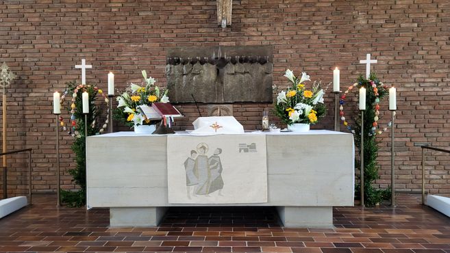 Kirchenaltar mit Blumen, Kerzen und einem Kreuz, umgeben von Ziegelwänden.