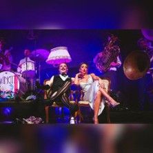 Eine Band auf der Bühne mit zwei Personen im Vordergrund, die auf Stühlen sitzen. Im Hintergrund sind Musiker mit Instrumenten zu sehen.