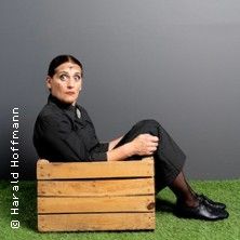 Eine Frau in schwarzer Kleidung sitzt in einer Holzkiste auf grünem Kunstrasen vor grauem Hintergrund.