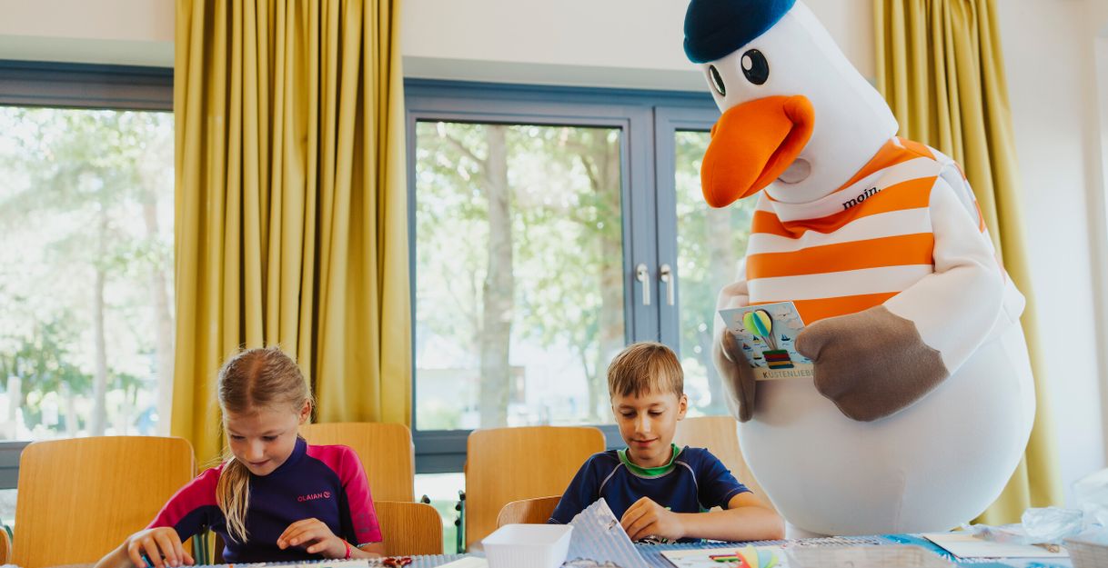 Zwei Kinder basteln an einem Tisch, während ein großes Möwenmaskottchen daneben steht.