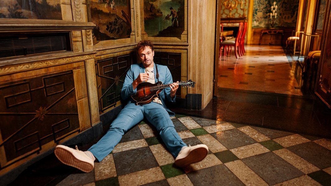 Person in blauem Anzug sitzt auf dem Boden mit Mandoline in einem dekorativen Raum.