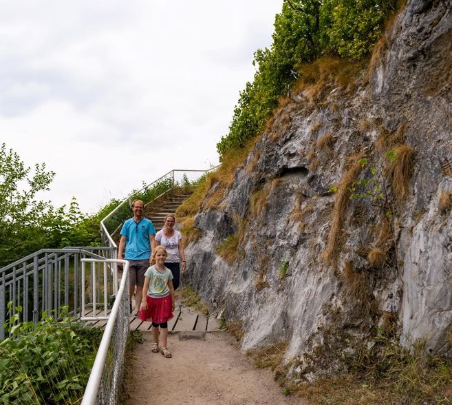 A family is walking along a path beside a steep rock face. In the background, there are stairs and a lot of green foliage visible.