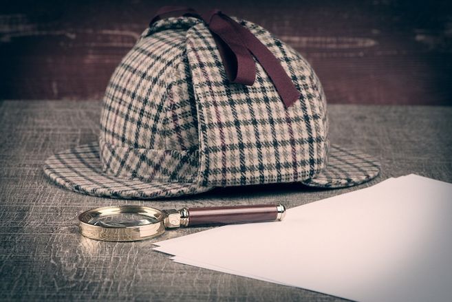 Eine karierte Detektivmütze, eine Lupe und weiße Papierblätter auf einem Tisch.