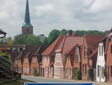 A picturesque street with historic houses and a church tower in the background. The sky is cloudy and the atmosphere is calm.