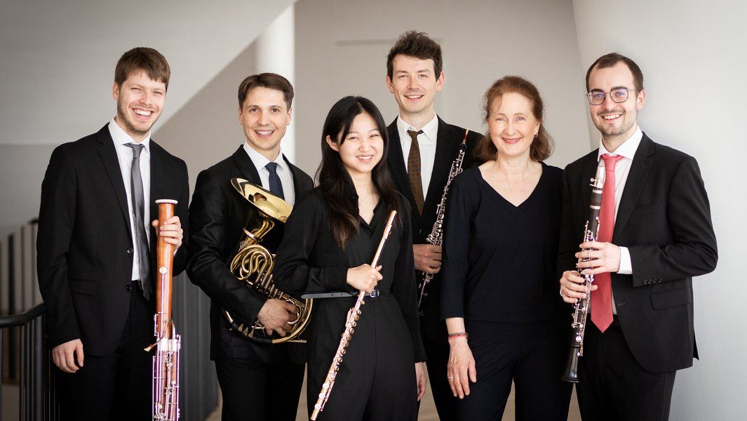 Eine Gruppe von sechs Musikern in formeller Kleidung mit verschiedenen Blasinstrumenten posiert für ein Foto.