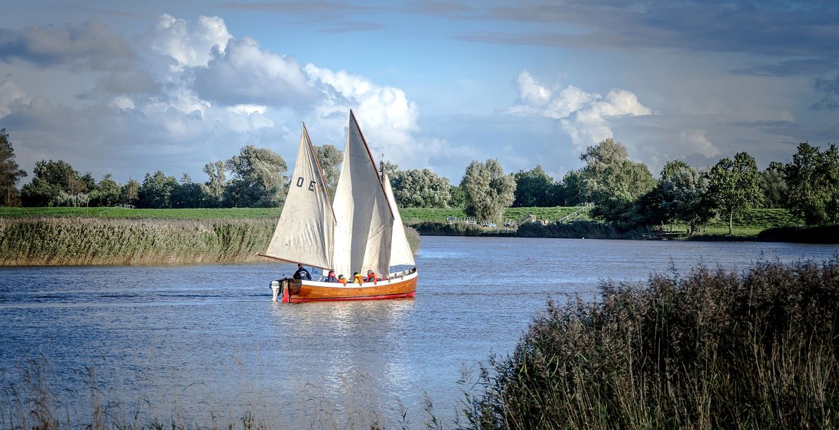 Ein Segelboot mit weißen Segeln fährt auf einem Fluss, umgeben von grüner Landschaft und blauem Himmel.