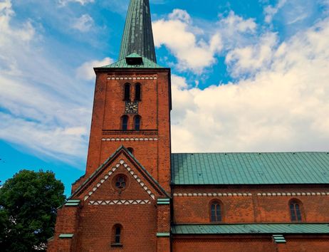A tall brick church with a pointed tower and a green roof. The sky is blue with some clouds.