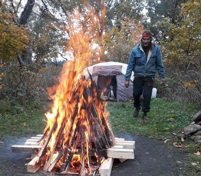 Ein Mann steht neben einem großen Lagerfeuer im Freien, im Hintergrund ist eine Schwitzhütte zu sehen.