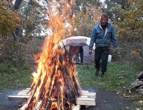 Ein Mann steht neben einem großen Lagerfeuer im Freien, im Hintergrund ist eine Schwitzhütte zu sehen.