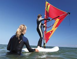 Zwei Personen beim Windsurfunterricht im Meer, eine steht auf dem Brett, die andere hält es fest.
