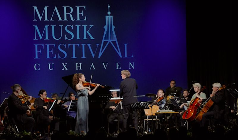 Ein Orchester spielt auf der Bühne des Mare Musik Festivals in Cuxhaven. Eine Geigerin steht im Vordergrund, während ein Dirigent das Ensemble leitet.