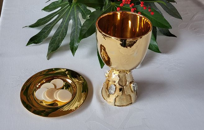 Goldener Kelch und Patene mit Hostien auf einem weißen Altartuch, umgeben von grünen Blättern und roten Beeren.
