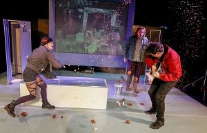 Theaterszene mit drei Personen auf der Bühne, umgeben von Requisiten und Projektionen.