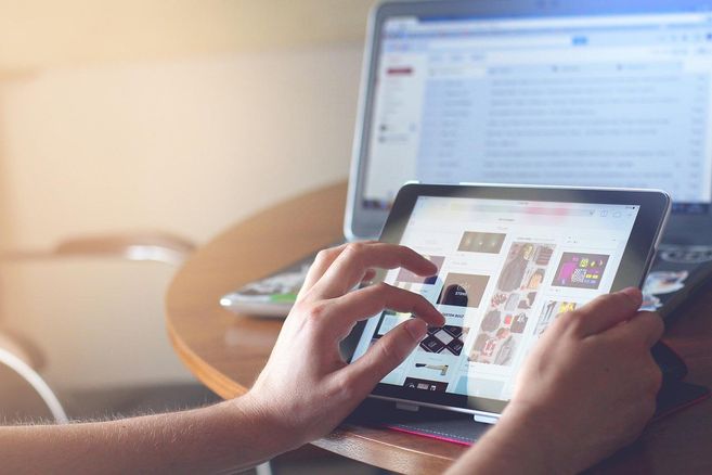 Person nutzt ein Tablet mit einem Laptop im Hintergrund auf einem Tisch.