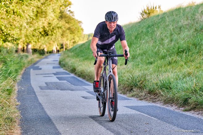 Ein Mann auf einem Rennrad fährt auf einem schmalen, asphaltierten Weg durch eine grüne Landschaft.